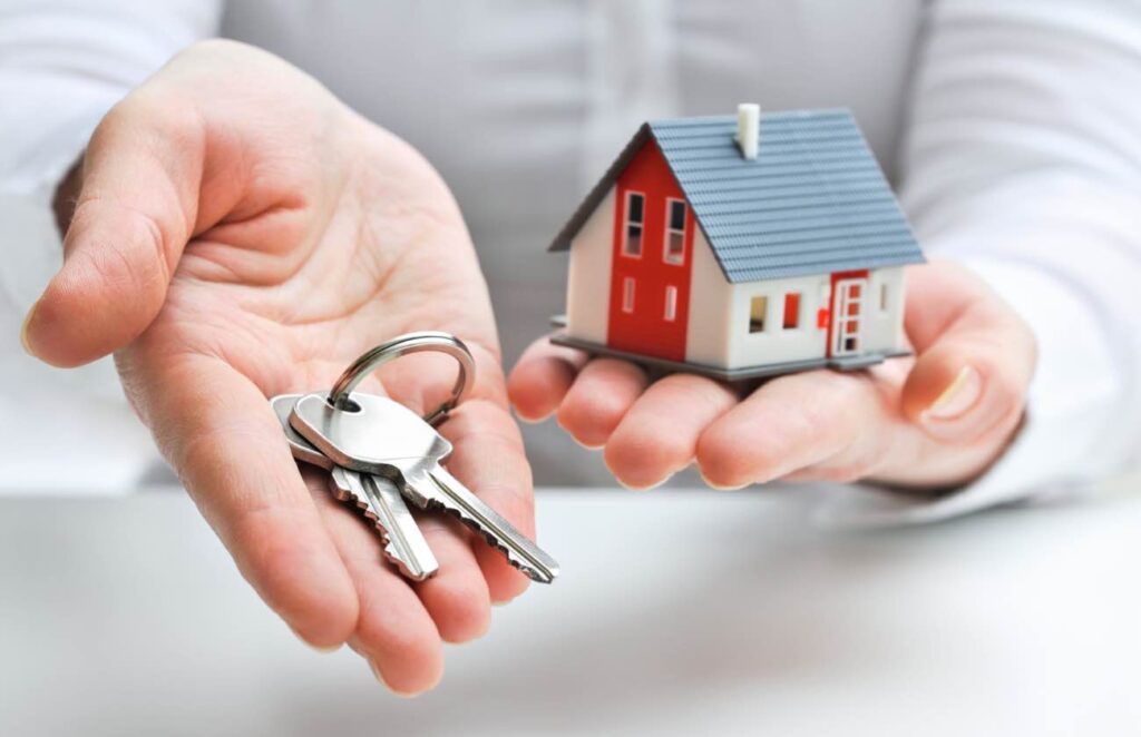 A person handing over a set of keys while holding a small model of a family home.